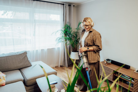 Smiling middle aged woman wiggling with uplifting music in headphones while getting floor clean with cordless vacuum. Housewife having fun while doing household chores. Housekeeping, home cleaningの写真素材