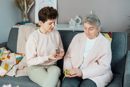 Middle aged daughter taking care of old mother, giving pills or vitamin supplements and glass of water helping her mom taking medicine on time. Elderly healthcare and support. Parenting love, care.の写真素材