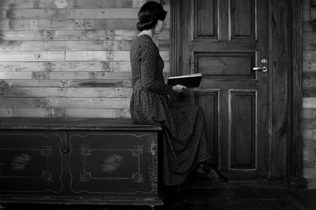 Beautiful woman sits in profile on a black chest and reads a book on a wooden backgroundの写真素材