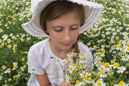 Portrait of a beautiful girl in a white dress and a hat inhaling the fragrance of daisy flowers on a meadowの写真素材