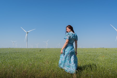 Beautiful woman in blue dress on green field with wind generator turbines and blue sky background