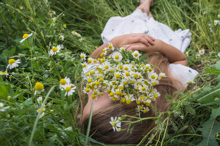 Beautiful little girl in a white dress is lying on a green meadow with a bouquet of daisiesの写真素材