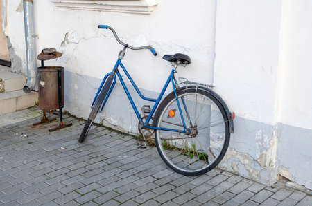 Old rarity blue bicycle against the background of a white ragged wall and a drain pipe and trash can with cigarette buttsの写真素材