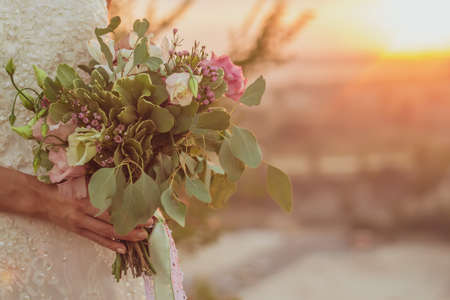Bride in white dress holds wedding bouquet against the background of the sunset. Close-up. Blurred background. Selective focus.の写真素材