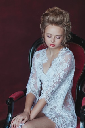shy and Young beautiful bride in elegant white robe sitting on a chair in a marsala interior.の写真素材