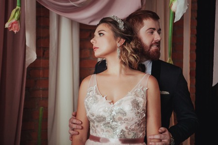 girl and bearded red man standing near the ceremonial arches with fabrics, fresh flowers on a background of brick wall. couple looking in different directions.の写真素材