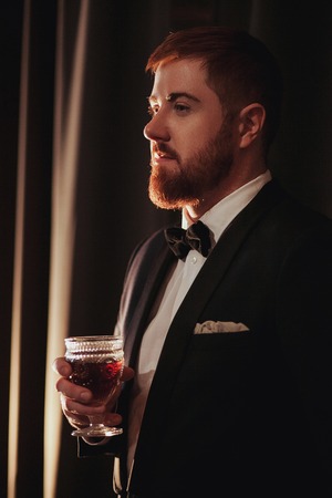 Young ginger bearded man holding a vintage glass with red wine against the light, black on background. view profile.の写真素材