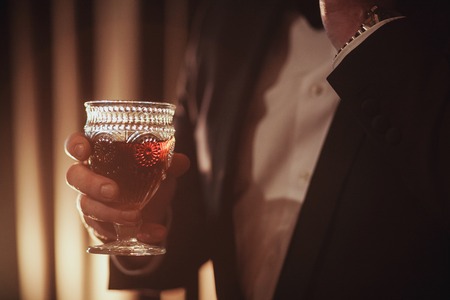 Young ginger bearded man holding a vintage glass with red wine against the light, black on background.の写真素材