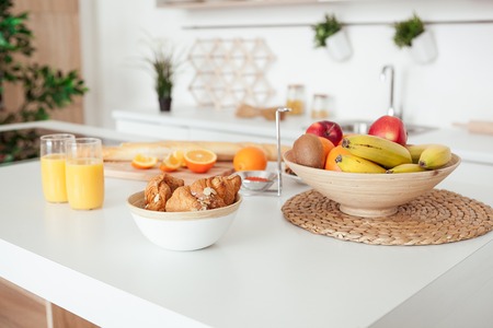 Plate of tasty cookies with chocolate crumbs, croissant, baget, bananas, apples, persimmon, orange and orange juice on white tableの写真素材