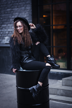 Portrait of a beautiful girl with brown eyes in glasses in a black hat and coat with fur sits on a barrel in the cityscape and looking down. The girl is like Harry Potterの写真素材