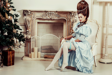 Indoors shot in the Marie Antoinette style. A young luxurious girl in a lush blue retro dress with a high hairstyle sits in a chair. Woman tired of luxuryの写真素材