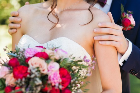 Handsome groom in dark blue suit gently holding beautiful bride with bridal bouquet of roses and peonies by the shouldersの写真素材
