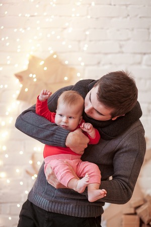 Dad wears knitted sweater and scarf holds on his hands a small son in red T-shirt standing in a...