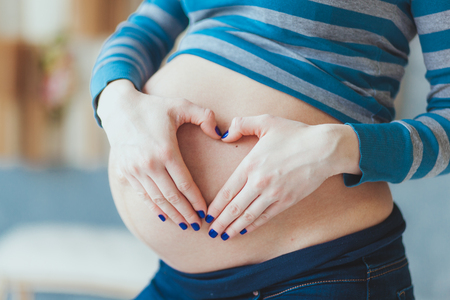 expectation concept - close up of happy pregnant woman making heart shape hand sign on her bare tummyの写真素材