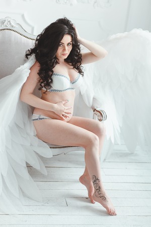 Beautiful young pregnant girl with tattoo and big white angel wings in white studio.の写真素材
