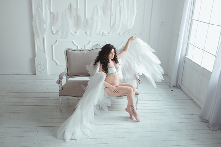 Beautiful young pregnant girl with big angel wings in white studio.の写真素材