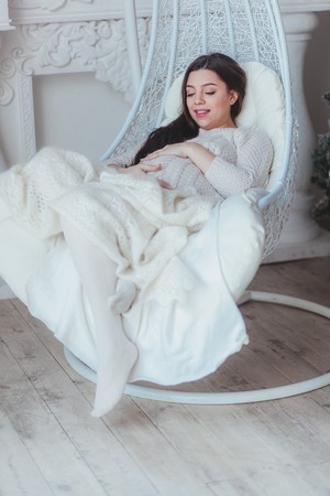 Beautiful pregnant woman sitting on wicker cocoon chair at homeの写真素材