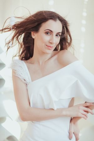 Beautiful tender woman in white dress posing by the background of white fabric christmas tree. Beauty, fashion. Haircare. Cosmetics.の写真素材