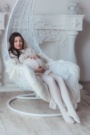Young pregnant woman in white knitted sweater sitting in armchair at home and touching her belly. Pregnancy and expectation conceptの写真素材
