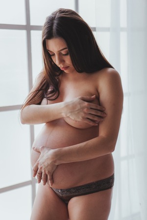 Pregnant caressing her nacked belly in front of window with curtain. Naked woman in lingerieの写真素材