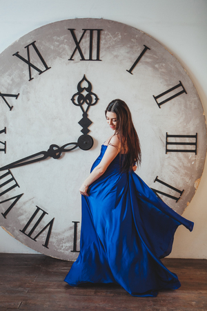 A pregnant woman in a beautiful blue long dress standing in front of huge clockの写真素材