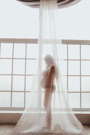 Nude beautiful young mom standing in front of window covered with chiffon curtain.の写真素材
