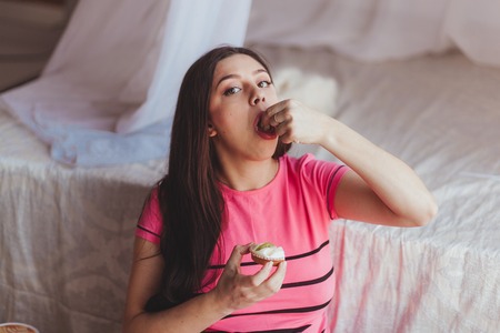 Attractive black hair pregnant woman eating cake with hands. Close-up studio portrait in bedroomの写真素材