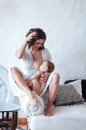 A young woman breast-feeding baby sitting on sofa with white rustic wall backgroundの写真素材