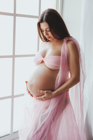 Close-up portrait of tender pregnant beauty young woman standing in front of the window and touching her belly at home. Loft minimalistic interiorの写真素材