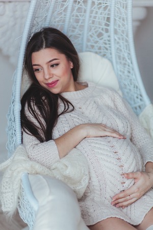 Young pregnant woman in white knitted sweater sitting in armchair at home and touching her belly. Pregnancy and expectation conceptの写真素材