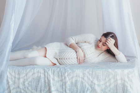 Pregnant beautifull woman lying in canopy bed. Wearing white knitted sweater and stockings.の写真素材