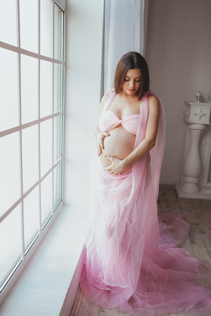Portrait of tender pregnant beauty young woman standing in front of the window and touching her belly at home. Loft minimalistic interiorの写真素材