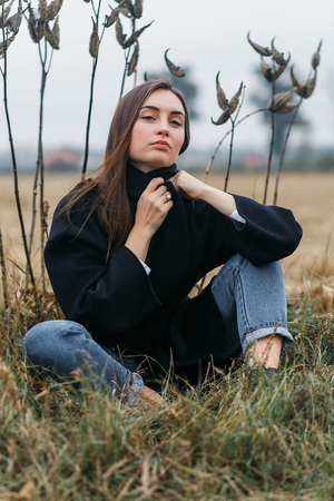 sad young woman sitting in long dry autumn grass seeking for solitude and peaceful silence from the city, cloudy weatherの写真素材