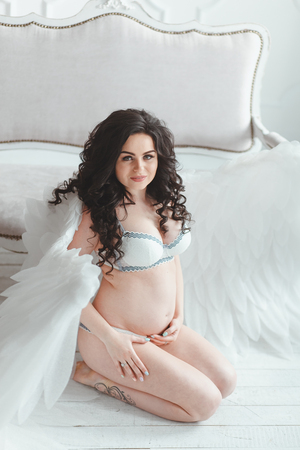 Beautiful young pregnant girl with big angel wings in white studio.の写真素材