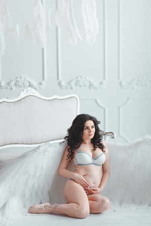 Beautiful young pregnant girl with big angel wings in white studio. Sitting on the floor near vintage sofaの写真素材