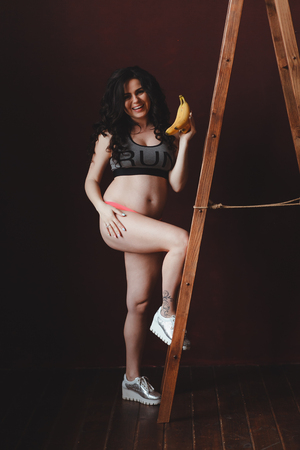 pregnant young woman fit looking, holding bananas at the ladder in loft studioの写真素材