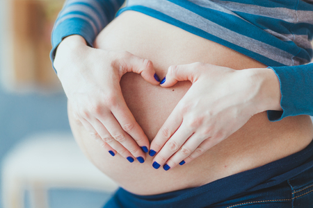 expectation concept - close up of happy pregnant woman making heart shape hand sign on her bare tummyの写真素材