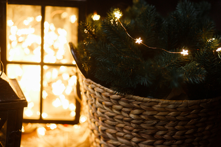 Christmas decor, wooden basket full of fir branches, lantern with led garland on background blured in the dark.の写真素材