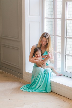 Happy cheerful mother playing with baby hugging near window.の写真素材
