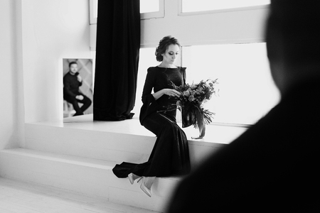 The bride in black dress. Reflecting in the mirror. White studio room with windows. Black and white shot with blurred groom silhouetteの写真素材