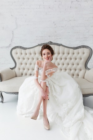 Beautiful, elegant bride sitting on vintage sofa in white nterior with brick wall backgroundの写真素材