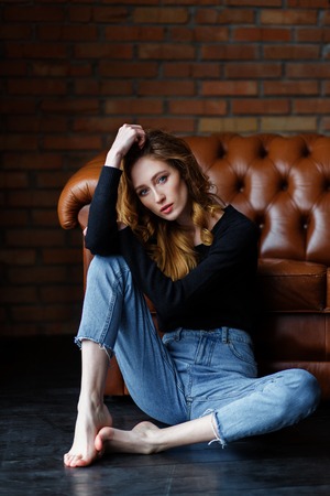 Portrait of a casual woman sitting on the floor and looking to camera on brown leather sofa and brick wall background.の写真素材