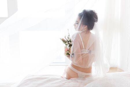 Tender beautiful bride in lingerie and veil sitting on bed in front of windowの写真素材