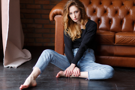 Portrait of elegant woman sitting on floor wearing a blue jeans and fur black sweater next to leather brown sofaの写真素材
