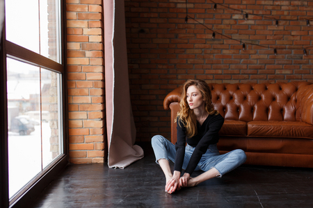 Loft style interior. sexual woman sits on the floor next to leather sofa in an expressive pose, looks at the window. Urban apartmentの写真素材