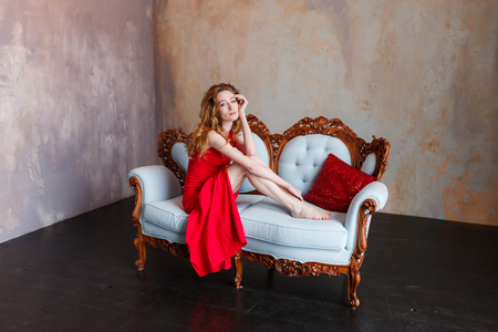 Luxurious redhead woman in a red dress on white classic couch in minimalistic loft interiorの写真素材
