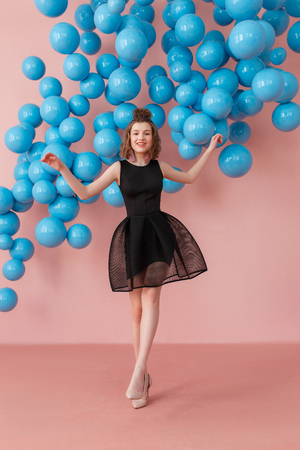Young woman in black dress standing in pink powder studio room with blue shiny balls on background. Modern punchy colorsの写真素材