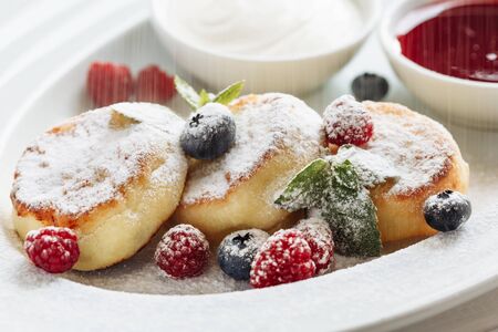 Close cheese pancakes, syrniki, curd fritters with fresh raspberries on white plate background. Decorated with powdered sugar falling down.の写真素材