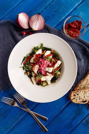 Fresh salad with sun-dried tomatoes, olives, arugula and onion. Top view decorated on blue tableの写真素材