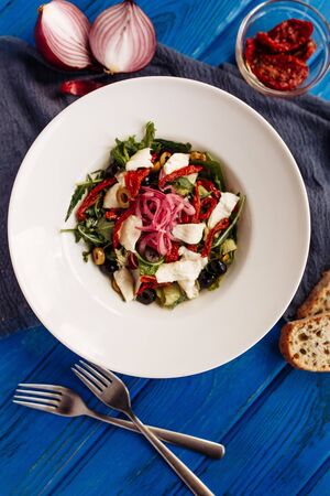 Fresh salad with sun-dried tomatoes, olives, arugula and onion. Top view decorated on blue tableの写真素材